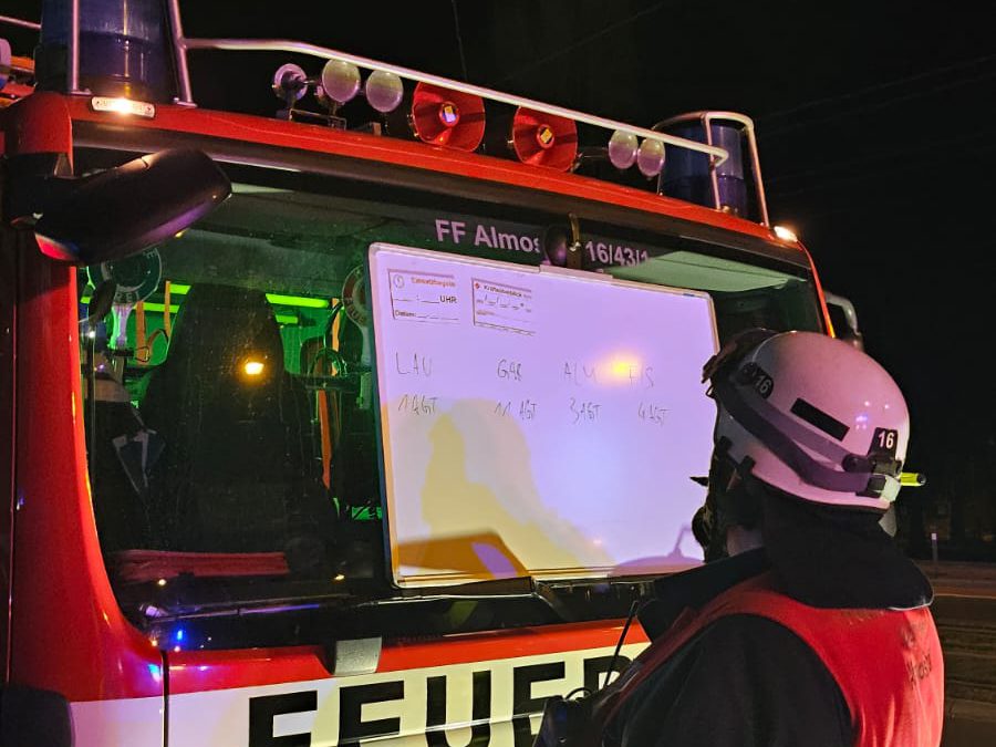 Ein Feuerwehrmann mit Löschzugführerweste steht vor einem Einsatzfahrzeug der FF Almoshof. An der Frontscheibe ist ein Whiteboard mit Lagedaten und Atemschutztrupps befestigt.