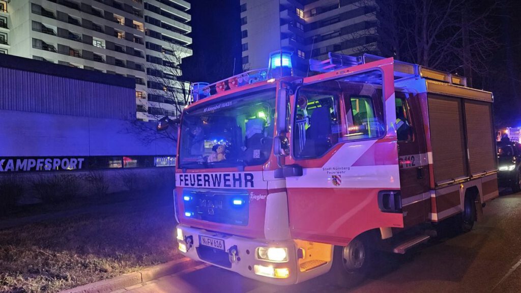 Einsatzfahrzeug der Freiwilligen Feuerwehr Almoshof bei einem nächtlichen Brandeinsatz vor einem Hochhaus in Nürnberg. Blaulicht leuchtet, mehrere Einsatzkräfte im Fahrzeug.