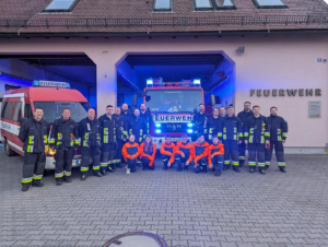 Gruppenfoto der Feuerwehr Almoshof vor dem Gerätehaus: Einsatzkräfte und Jugendfeuerwehr stehen und knien gemeinsam vor zwei Fahrzeugen mit eingeschaltetem Blaulicht.