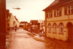 Aufnahme vom Hochwasser 1979 nach dem Dammbruch in Katzwang. Ein Hubschrauber fliegt über eine überflutete Straße mit beschädigten Gebäuden im Hintergrund.